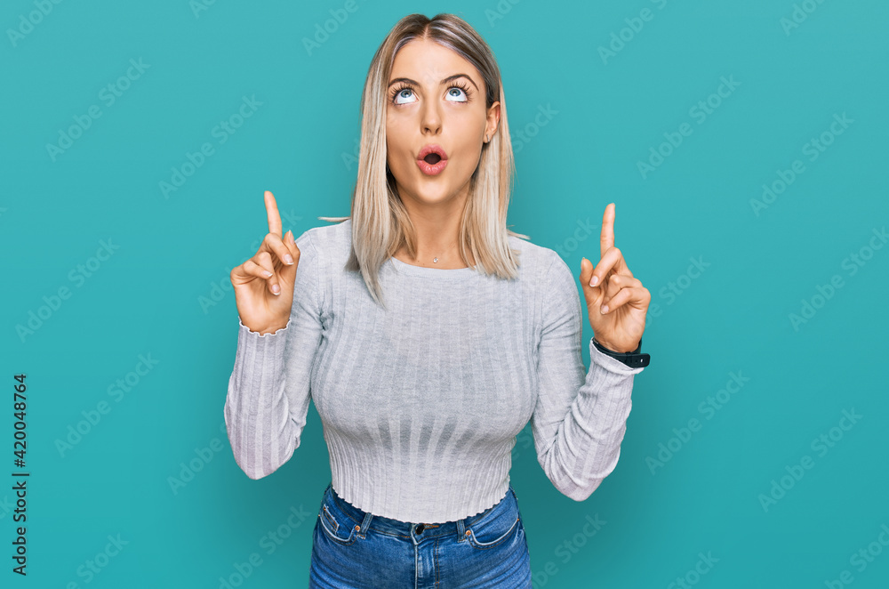 Beautiful blonde woman wearing casual clothes amazed and surprised looking up and pointing with fingers and raised arms. Beautiful blonde woman wearing casual clothes amazed and surprised looking up and pointing with fingers and raised arms.