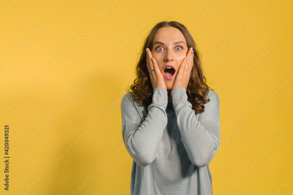 Shocked and amazed young attractive woman looks at the camera isolated on yellow, a curly caucasian girl screams surprised, holding cheeks, incredible sales or deal