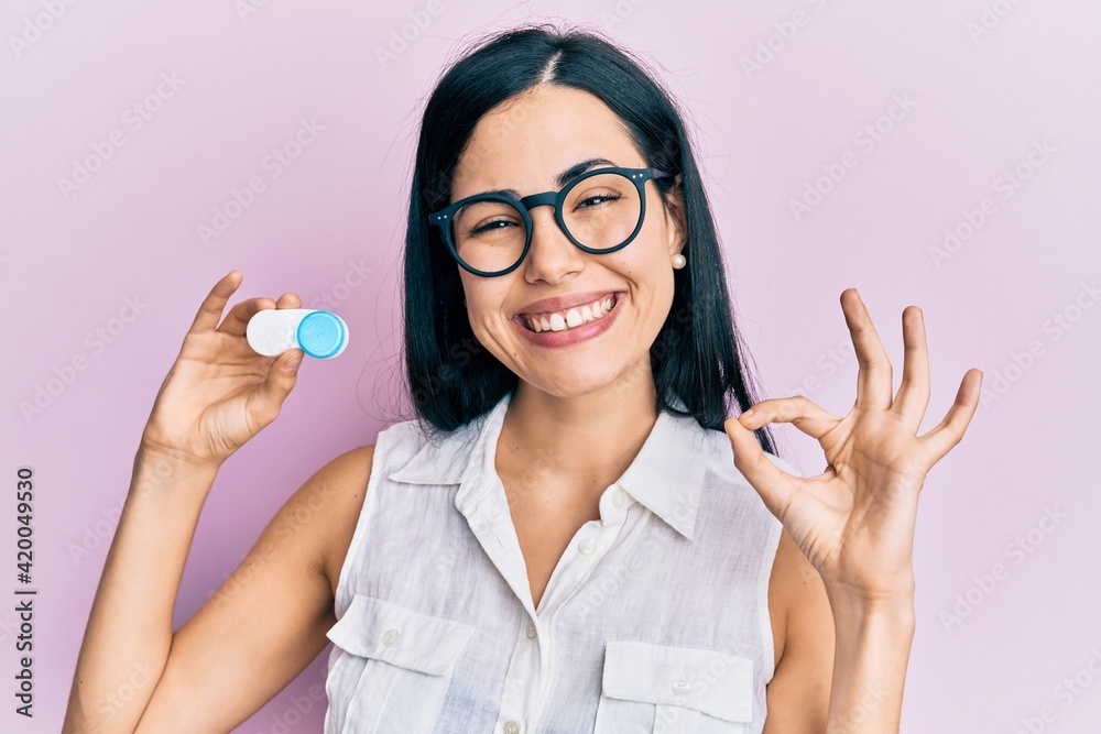 Beautiful young woman holding glasses and contact lenses doing ok sign with fingers, smiling friendly gesturing excellent symbol Beautiful young woman holding glasses and contact lenses doing ok sign with fingers, smiling friendly gesturing excellent symbol