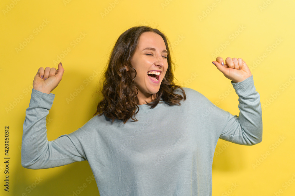 Joyful and happy young woman dancing alone, enjoying good day, a female with cheerful face expression in blue jumper has fun, studio shot isolated on yellow Joyful and happy young woman dancing alone, enjoying good day, a female with cheerful face expression in blue jumper has fun, studio shot isolated on yellow