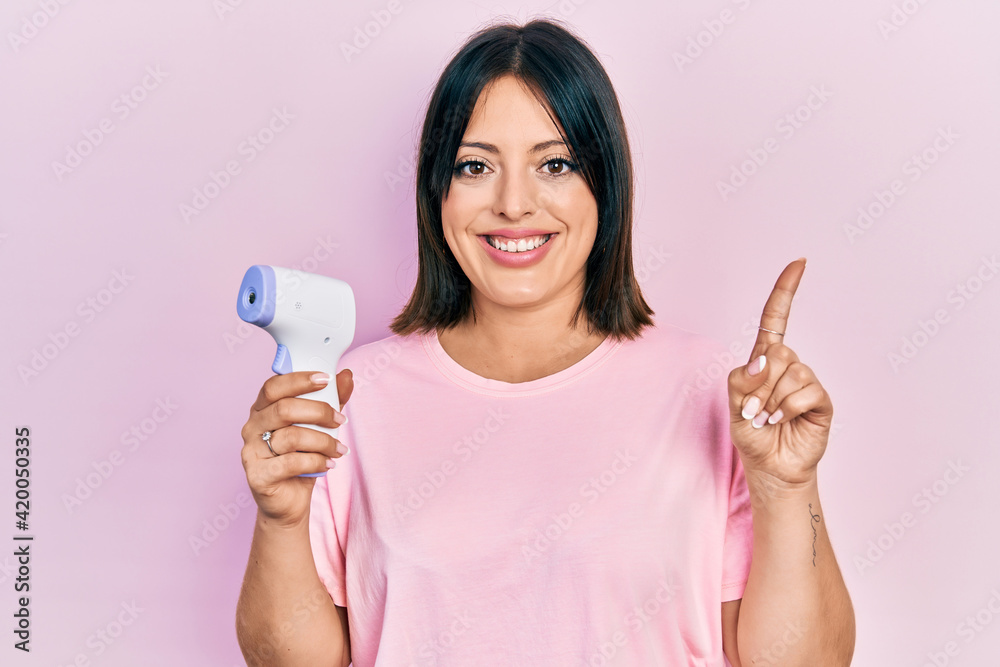 Young hispanic woman holding thermometer surprised with an idea or question pointing finger with happy face, number one Young hispanic woman holding thermometer surprised with an idea or question pointing finger with happy face, number one
