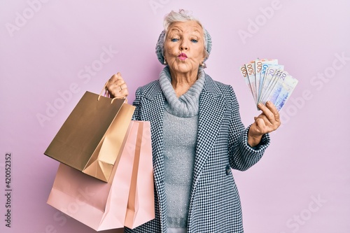 Senior grey-haired woman holding shopping bags and swedish krona banknotes looking at the camera blowing a kiss being lovely and sexy. love expression.