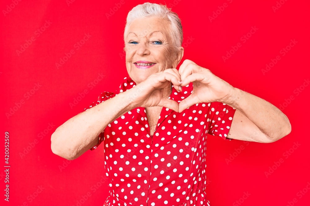Senior beautiful woman with blue eyes and grey hair wearing casual summer clothes over red background smiling in love doing heart symbol shape with hands. romantic concept. Senior beautiful woman with blue eyes and grey hair wearing casual summer clothes over red background smiling in love doing heart symbol shape with hands. romantic concept.