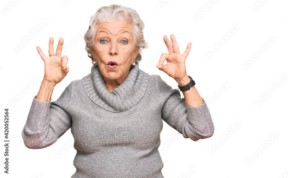 Senior grey-haired woman wearing casual winter sweater looking surprised and shocked doing ok approval symbol with fingers. crazy expression Senior grey-haired woman wearing casual winter sweater looking surprised and shocked doing ok approval symbol with fingers. crazy expression