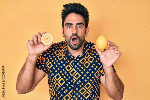 Handsome hispanic man with beard holding lemon afraid and shocked with surprise and amazed expression, fear and excited face.