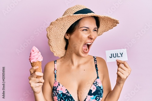 Young brunette woman with short hair holding ice cream and july paper angry and mad screaming frustrated and furious, shouting with anger. rage and aggressive concept.