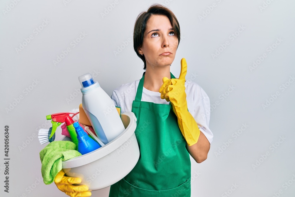 Young brunette woman with short hair wearing apron holding cleaning products pointing up looking sad and upset, indicating direction with fingers, unhappy and depressed. Young brunette woman with short hair wearing apron holding cleaning products pointing up looking sad and upset, indicating direction with fingers, unhappy and depressed.