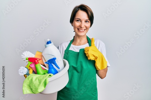 Young brunette woman with short hair wearing apron holding cleaning products doing happy thumbs up gesture with hand. approving expression looking at the camera showing success.