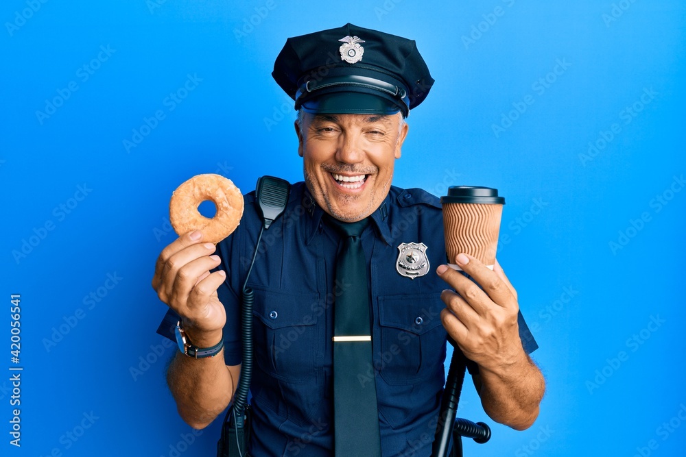 Handsome middle age mature police man eating donut and drinking coffee smiling and laughing hard out loud because funny crazy joke.