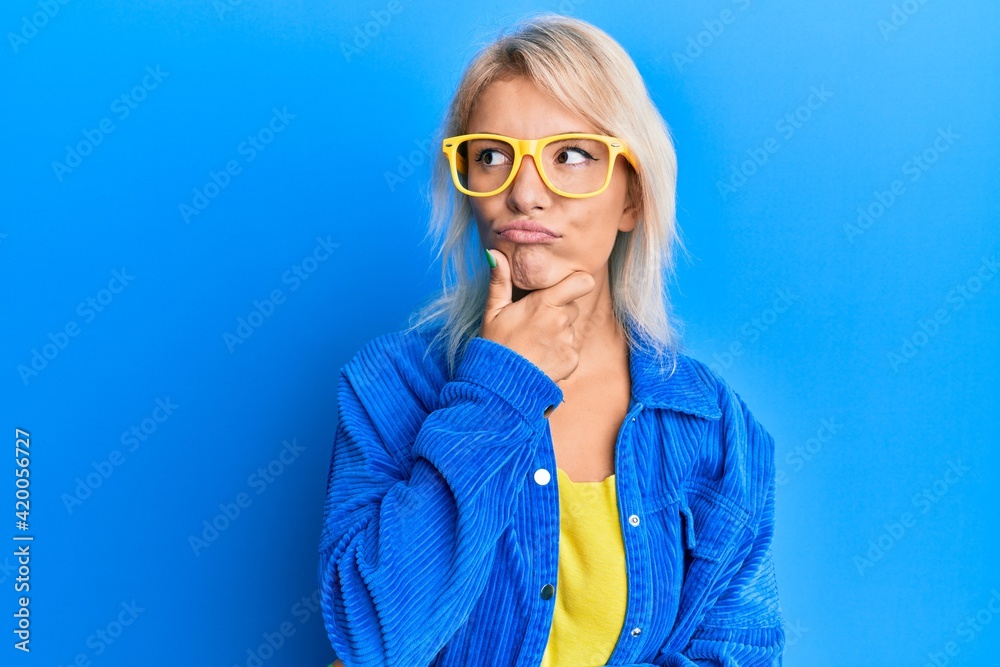 Young blonde girl wearing casual clothes and glasses serious face thinking about question with hand on chin, thoughtful about confusing idea Young blonde girl wearing casual clothes and glasses serious face thinking about question with hand on chin, thoughtful about confusing idea