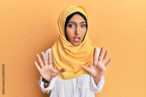 Young brunette arab woman wearing traditional islamic hijab scarf afraid and terrified with fear expression stop gesture with hands, shouting in shock. panic concept. © Krakenimages.com