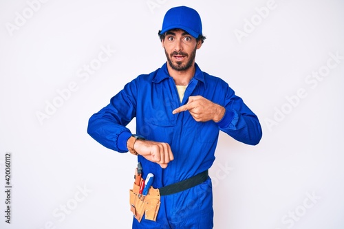 Handsome young man with curly hair and bear weaing handyman uniform in hurry pointing to watch time  impatience  upset and angry for deadline delay