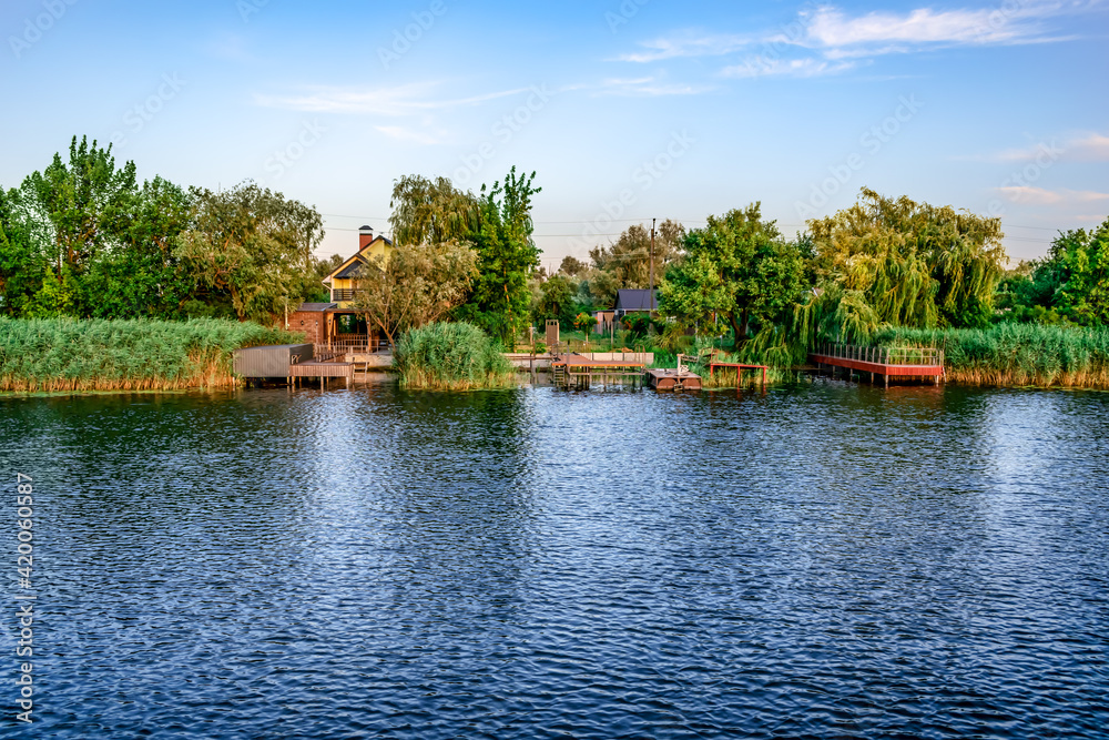 Residential houses with personal piers on Konka River coast in Kherson floodplains (Ukraine). Suburban area with buildings on the shore among green trees and reeds on the water surface background