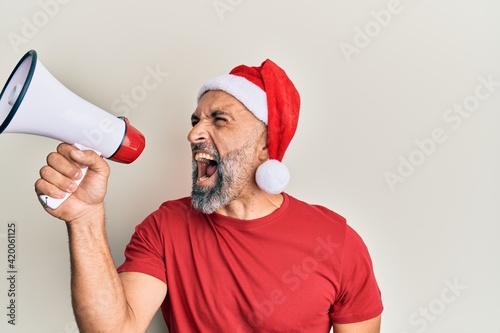 Middle age grey-haired man wearing santa claus hat screaming angry using megaphone over isolated white background.