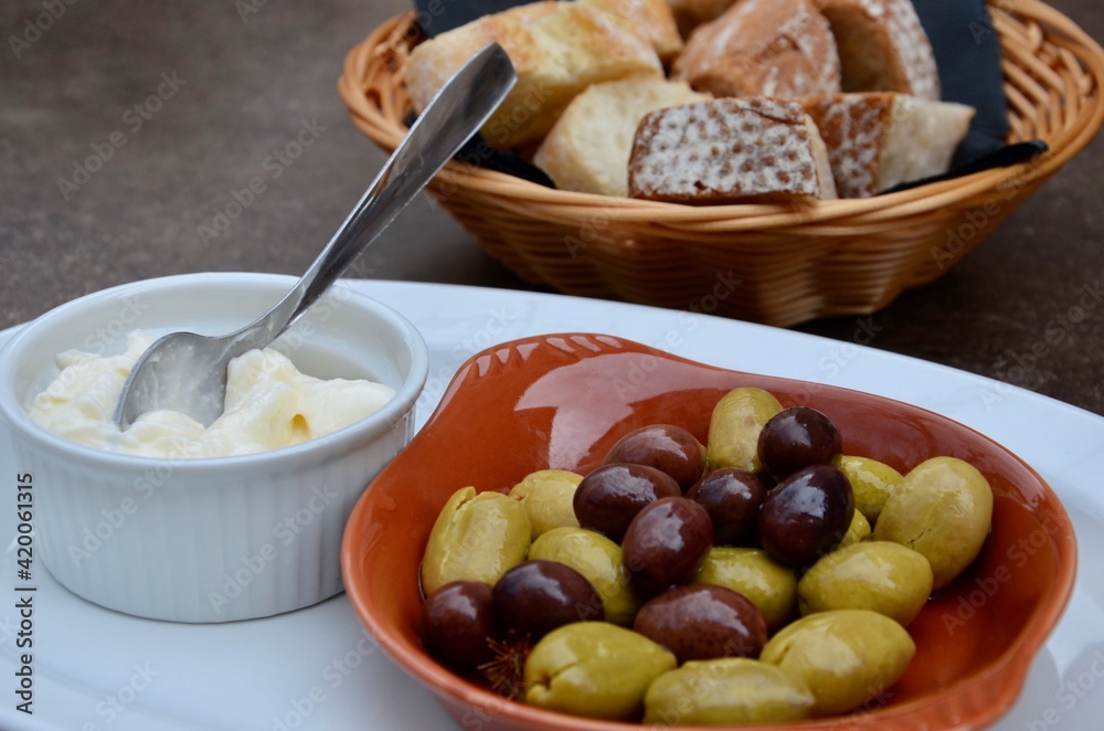 Green and black olives in a brown clay bowl, garlic cream (Allioli) next to it, basket with cut bread behind, a traditional snack on Balearic island Mallorca, Spain, blurred table background