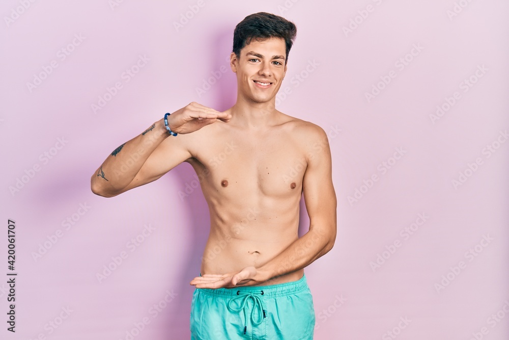 Fototapeta premium Young hispanic man wearing swimwear shirtless gesturing with hands showing big and large size sign, measure symbol. smiling looking at the camera. measuring concept.