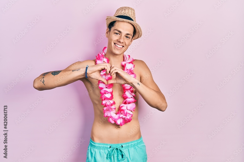 Young hispanic man wearing swimwear and summer hat smiling in love doing heart symbol shape with hands. romantic concept.