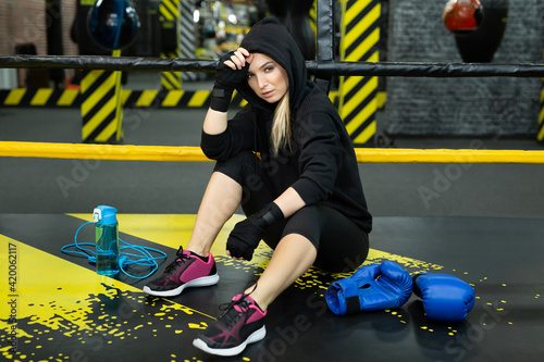 Young athletic girl in a black tracksuit sits in the ring and rests after a workout.