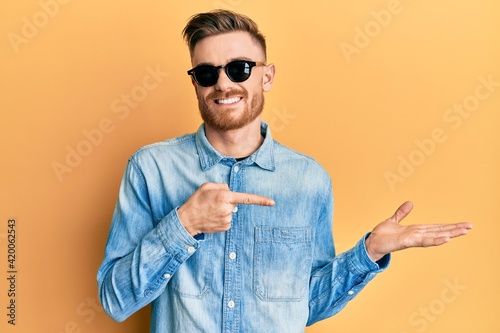 Young redhead man wearing stylish sunglasses amazed and smiling to the camera while presenting with hand and pointing with finger.