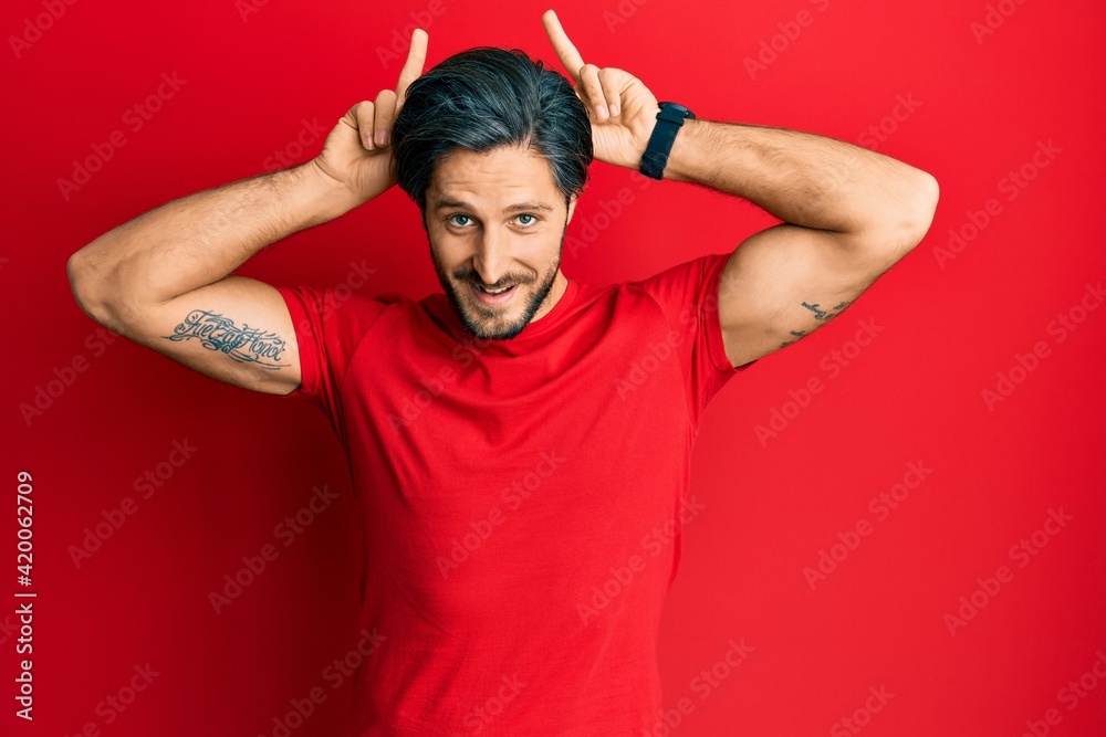 Fototapeta premium Young hispanic man wearing casual red t shirt posing funny and crazy with fingers on head as bunny ears, smiling cheerful