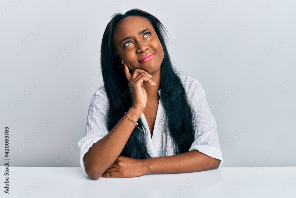 Young african woman wearing casual clothes sitting on the table thinking concentrated about doubt with finger on chin and looking up wondering Young african woman wearing casual clothes sitting on the table thinking concentrated about doubt with finger on chin and looking up wondering