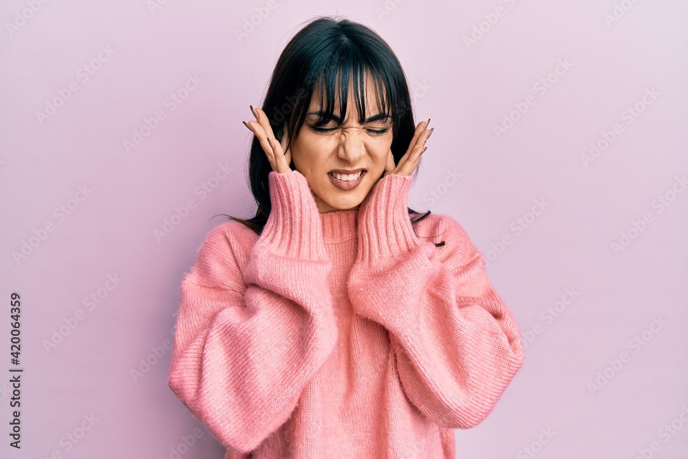 Young brunette woman with bangs wearing casual winter sweater covering ears with fingers with annoyed expression for the noise of loud music. deaf concept. Young brunette woman with bangs wearing casual winter sweater covering ears with fingers with annoyed expression for the noise of loud music. deaf concept.