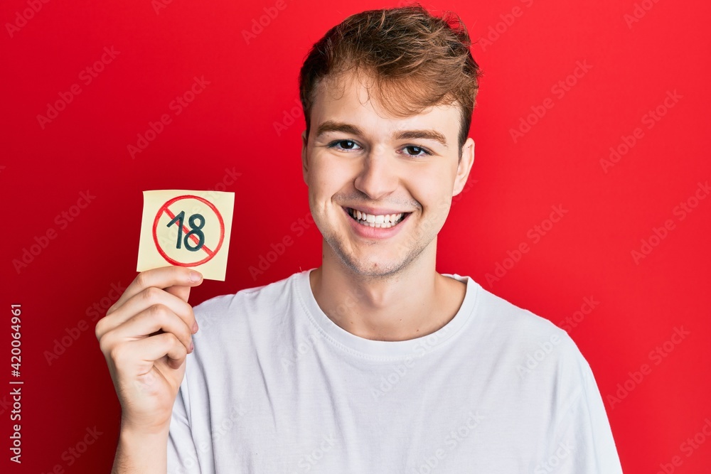 Young caucasian man holding under 18 prohibition sticker looking positive and happy standing and smiling with a confident smile showing teeth Young caucasian man holding under 18 prohibition sticker looking positive and happy standing and smiling with a confident smile showing teeth