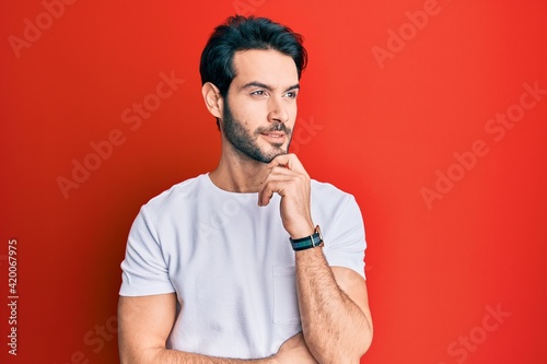 Young hispanic man wearing casual white tshirt with hand on chin thinking about question, pensive expression. smiling and thoughtful face. doubt concept.
