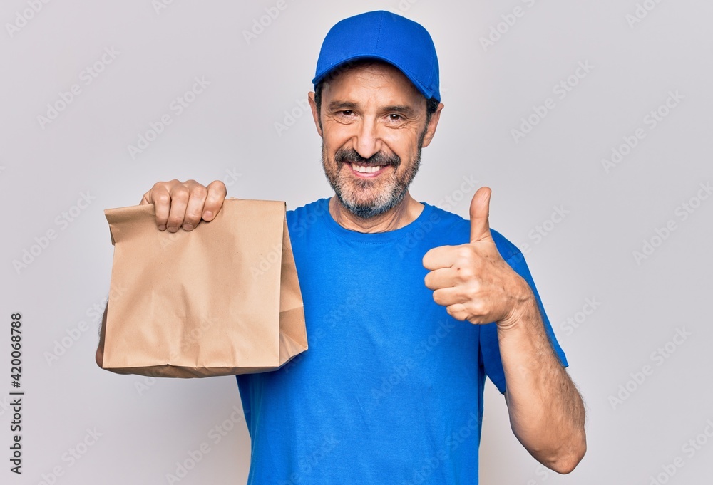 Middle age handsome delivery man wearing cap holding paper bag with takeaway food smiling happy and positive, thumb up doing excellent and approval sign Middle age handsome delivery man wearing cap holding paper bag with takeaway food smiling happy and positive, thumb up doing excellent and approval sign