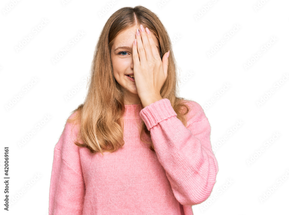 Beautiful young caucasian girl wearing casual winter sweater covering one eye with hand, confident smile on face and surprise emotion.
