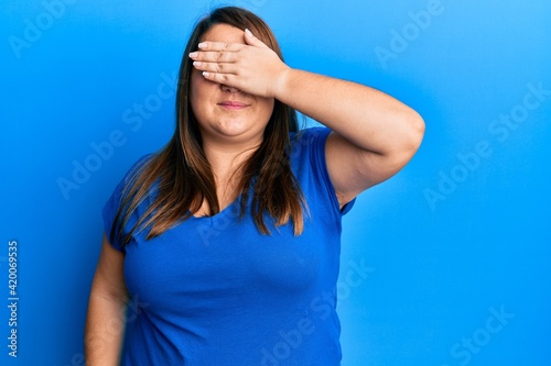 Beautiful brunette plus size woman wearing casual blue t shirt smiling and laughing with hand on face covering eyes for surprise. blind concept.