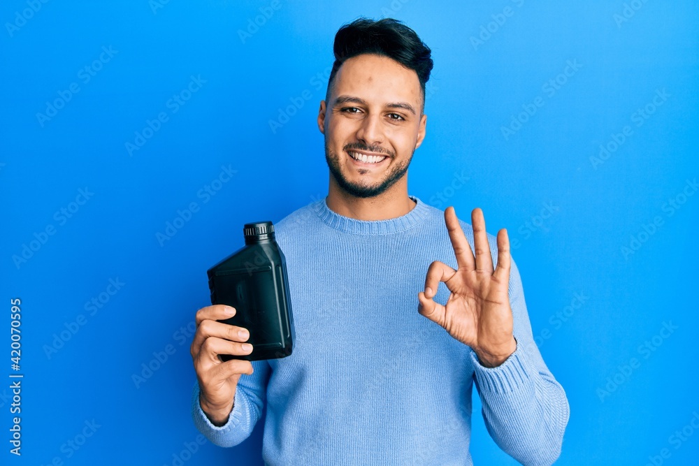 Young arab man holding motor oil bottle doing ok sign with fingers, smiling friendly gesturing excellent symbol Young arab man holding motor oil bottle doing ok sign with fingers, smiling friendly gesturing excellent symbol