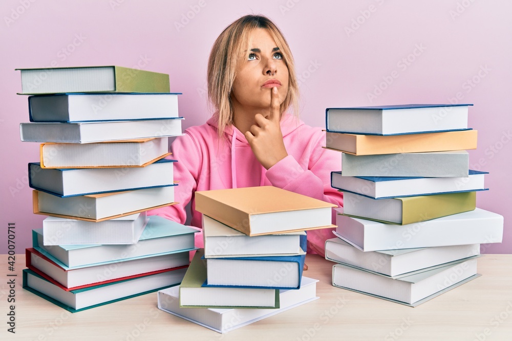 Young caucasian woman sitting on the table with books thinking concentrated about doubt with finger on chin and looking up wondering Young caucasian woman sitting on the table with books thinking concentrated about doubt with finger on chin and looking up wondering