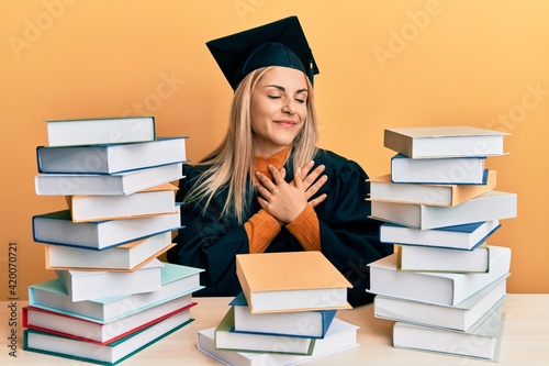 Young caucasian woman wearing graduation ceremony robe sitting on the table smiling with hands on chest with closed eyes and grateful gesture on face. health concept.