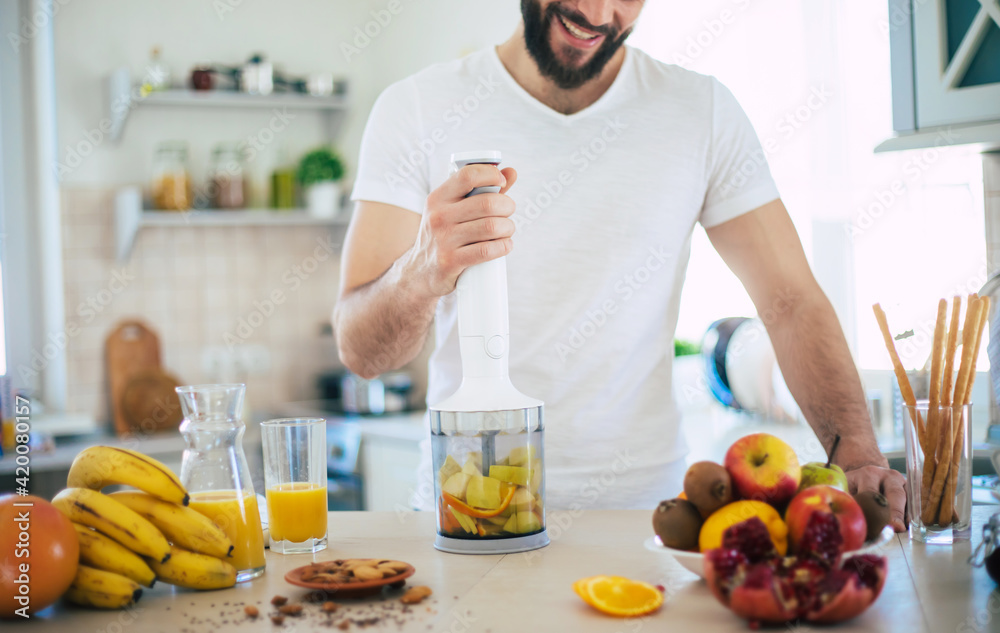 Handsome young sporty smiling man in the kitchen is preparing vegan healthy fruits salad and smoothie in a good mood - obrazy, fototapety, plakaty 