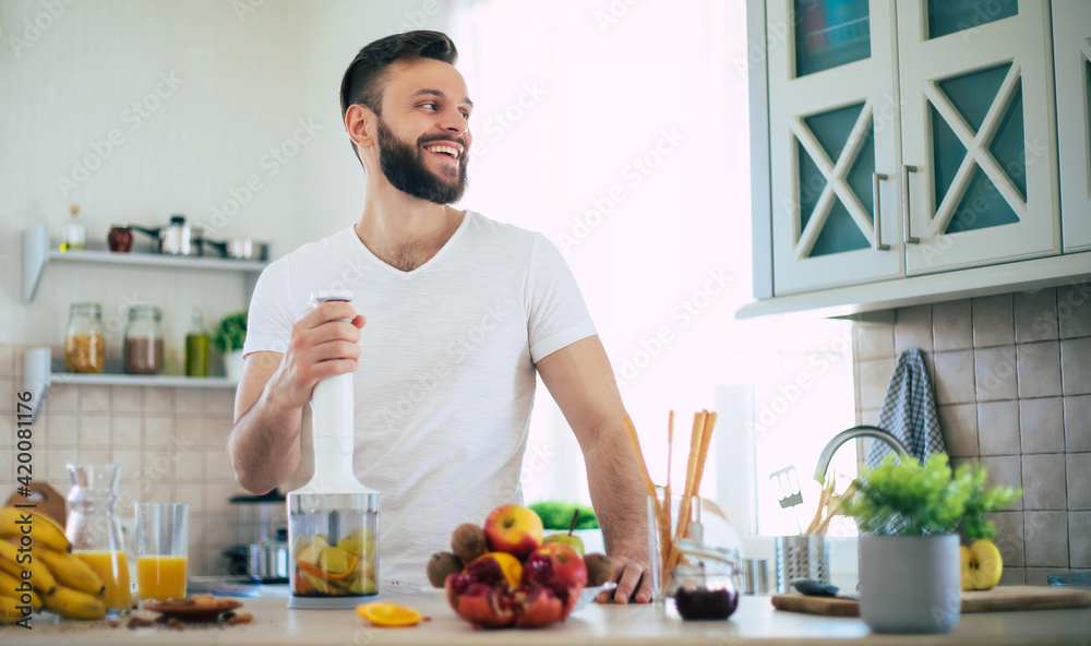 Handsome young sporty smiling man in the kitchen is preparing vegan healthy fruits salad and smoothie in a good mood Handsome young sporty smiling man in the kitchen is preparing vegan healthy fruits salad and smoothie in a good mood