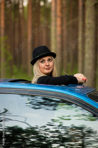 Fototapeta Naklejka Na Ścianę i Meble -  a blonde girl in a black hat looks out the hatch of a blue car, selective focus