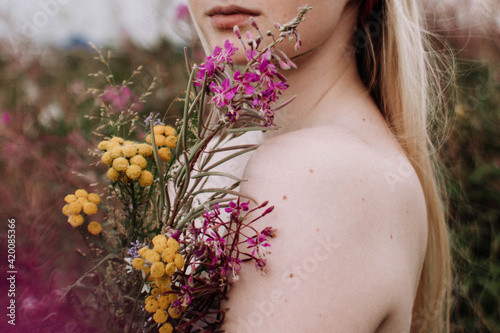 Young woman with naked sholders holding bouquet of yellow and purple wild flowers standing in green field at summer. Caucasian girl with blonde hair. Outdoors portrait in countryside.