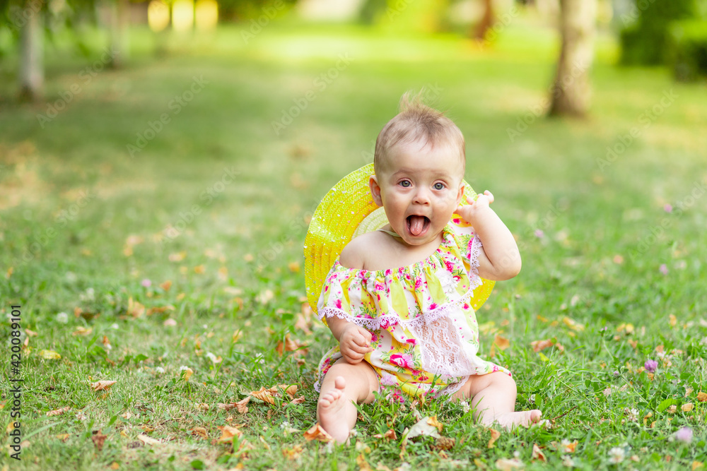 little baby girl 7 months old sitting on the green grass in a yellow dress and hat and holding her hand to her ear and showing her tongue, walking in the fresh air. Space for text little baby girl 7 months old sitting on the green grass in a yellow dress and hat and holding her hand to her ear and showing her tongue, walking in the fresh air. Space for text