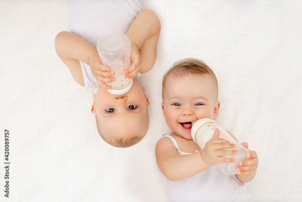 twin babies boy and girl with a bottle of milk on a white bed at home, baby food concept, place for text - obrazy, fototapety, plakaty 