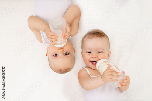 twin babies boy and girl with a bottle of milk on a white bed at home, baby food concept, place for text