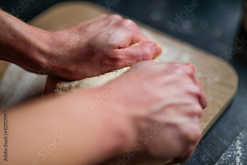 Close up and selective focus on the hands of an unidentifiable baker kneading bread dough on a wooden chopping board