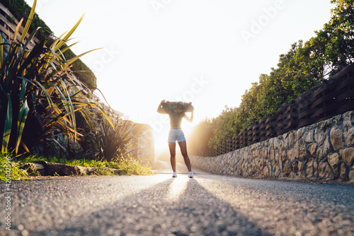 Beautiful caucasian female in trendy wear standing on asphalt road in sunrays holding board for skating on free time in evening, trendy casually dressed woman enjoying active hobby #420102596