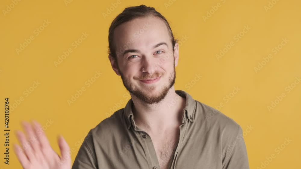Young attractive bearded man looking happy smiling and waving hello on camera over yellow background. Greeting expression Young attractive bearded man looking happy smiling and waving hello on camera over yellow background. Greeting expression