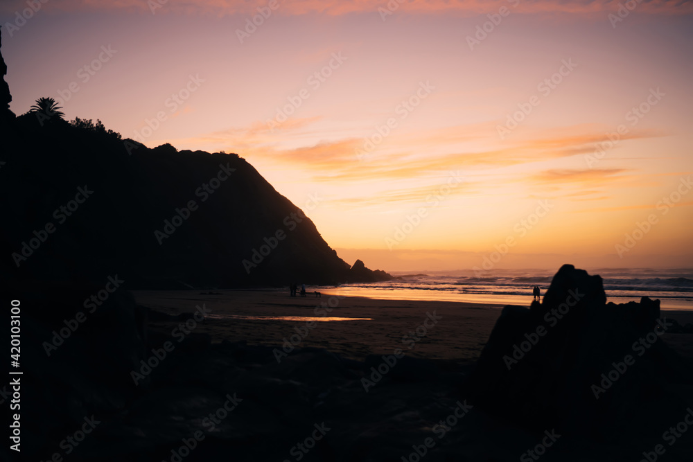 Dark evening view of ocean and high rocks on sandy shore with sunset on horizon, breathtaking scenery of marine and yellow sundown