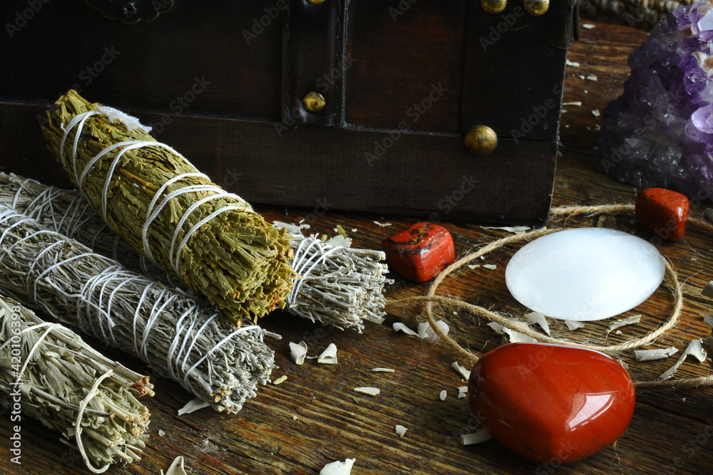 A close up image of a variety of sacred smudge bundles with selenite and red jasper crystals on a dark wooden table top. A close up image of a variety of sacred smudge bundles with selenite and red jasper crystals on a dark wooden table top.