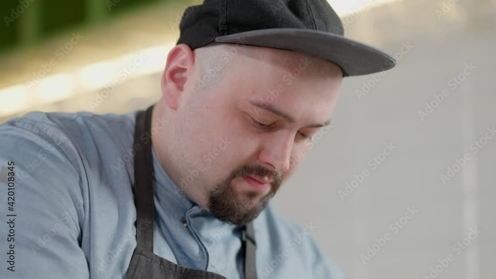 Headshot of concentrated Caucasian plus-size cook in hat and apron in kitchen indoors. Focused professional chef working in restaurant or cafe. Lifestyle and employment concept.