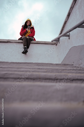 Young woman with mobile phone sitting on concrete parapet. Brunette in yellow hoodie and red jacket browsing smartphone in cloudy weather.