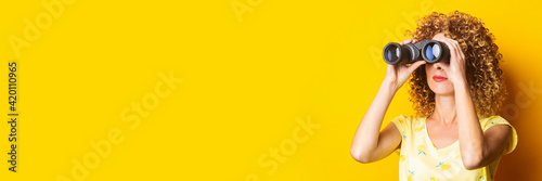 curly young woman looks through binoculars on a yellow background. Banner.