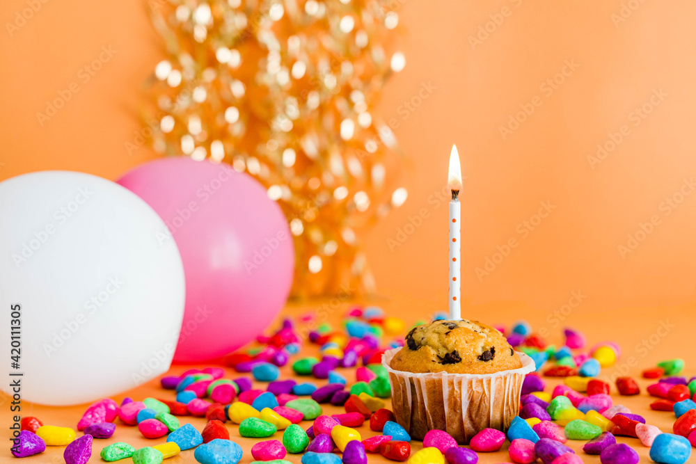 cartolina di buon compleanno con un muffin e una candelina accesa, due  palloncini colorati, pietre colorate e nastri dorati nello sfondo, su uno  sfondo arancione. vista frontale. buon compleanno Photos